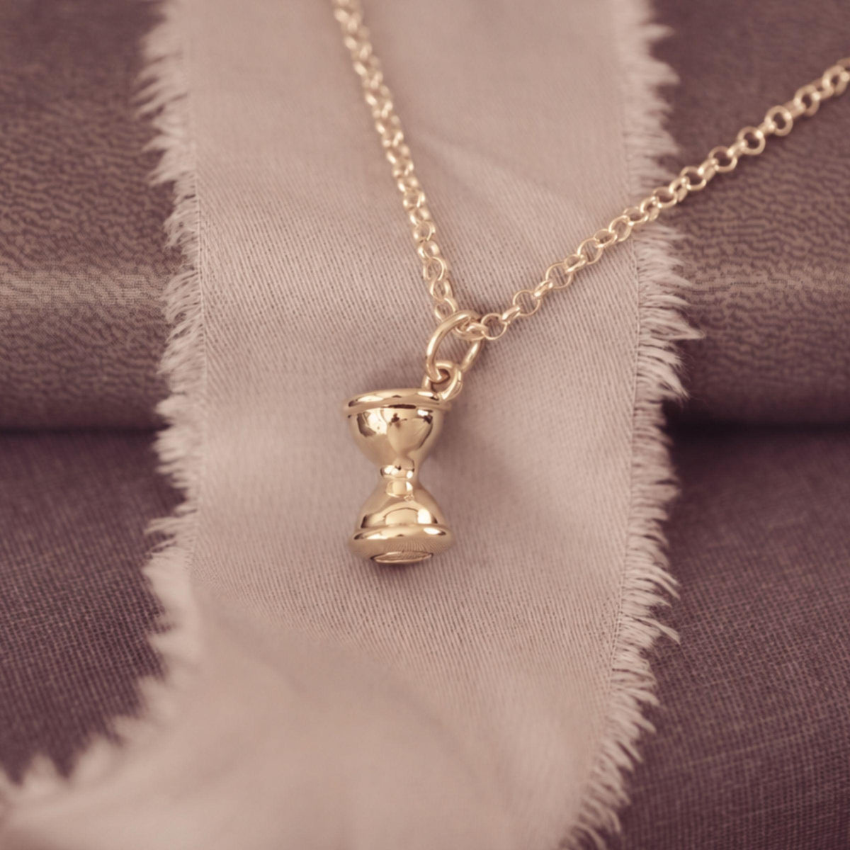 A gold-toned pendant necklace with a small hourglass charm, displayed on a textured fabric background with a frayed beige ribbon.