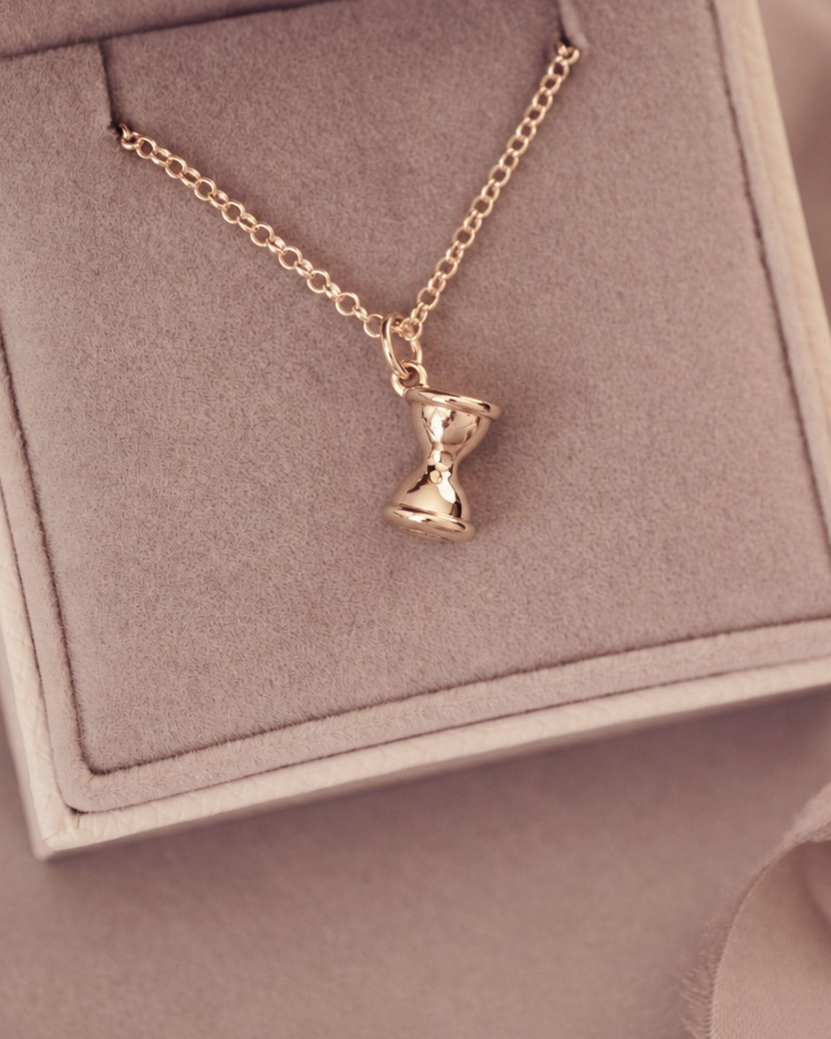 A gold chain necklace with a small hourglass pendant, displayed inside a beige jewelry box with a soft beige fabric background.