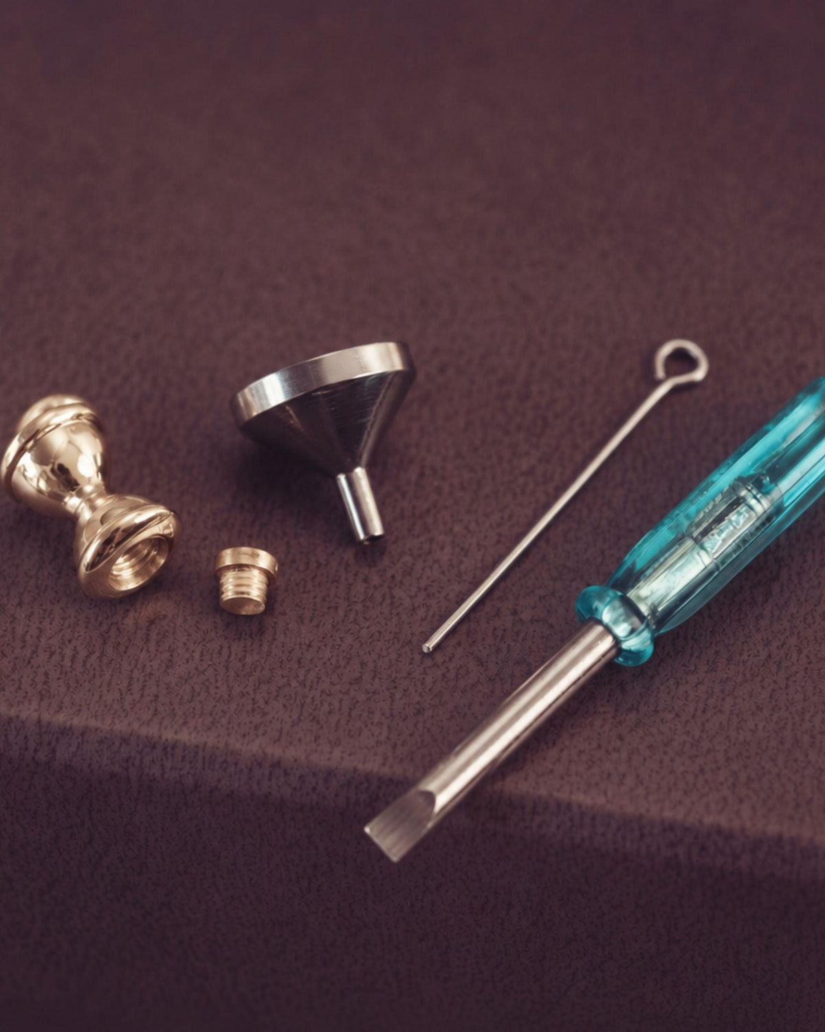 Jewellery tools - a metal funnel, a thin metal tool, and a blue-handled screwdriver on a dark brown textured surface.