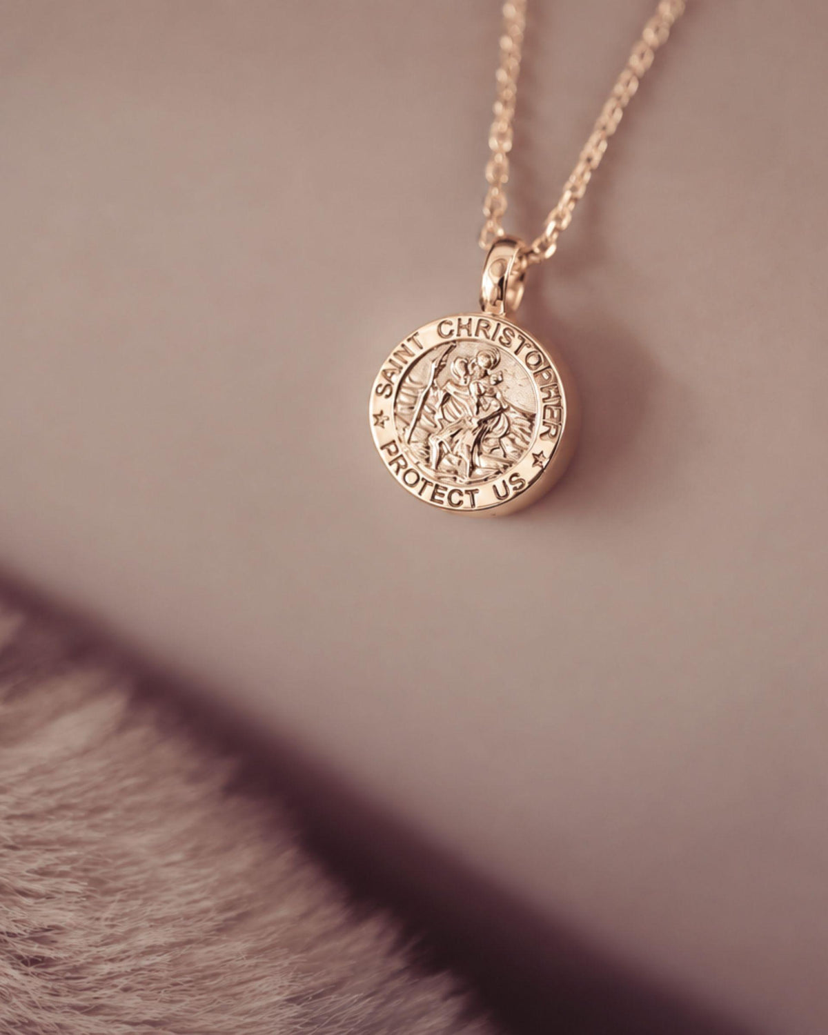 A close-up of a gold Saint Christopher pendant necklace resting on a neutral surface with a soft, out-of-focus textured prop in the foreground.
