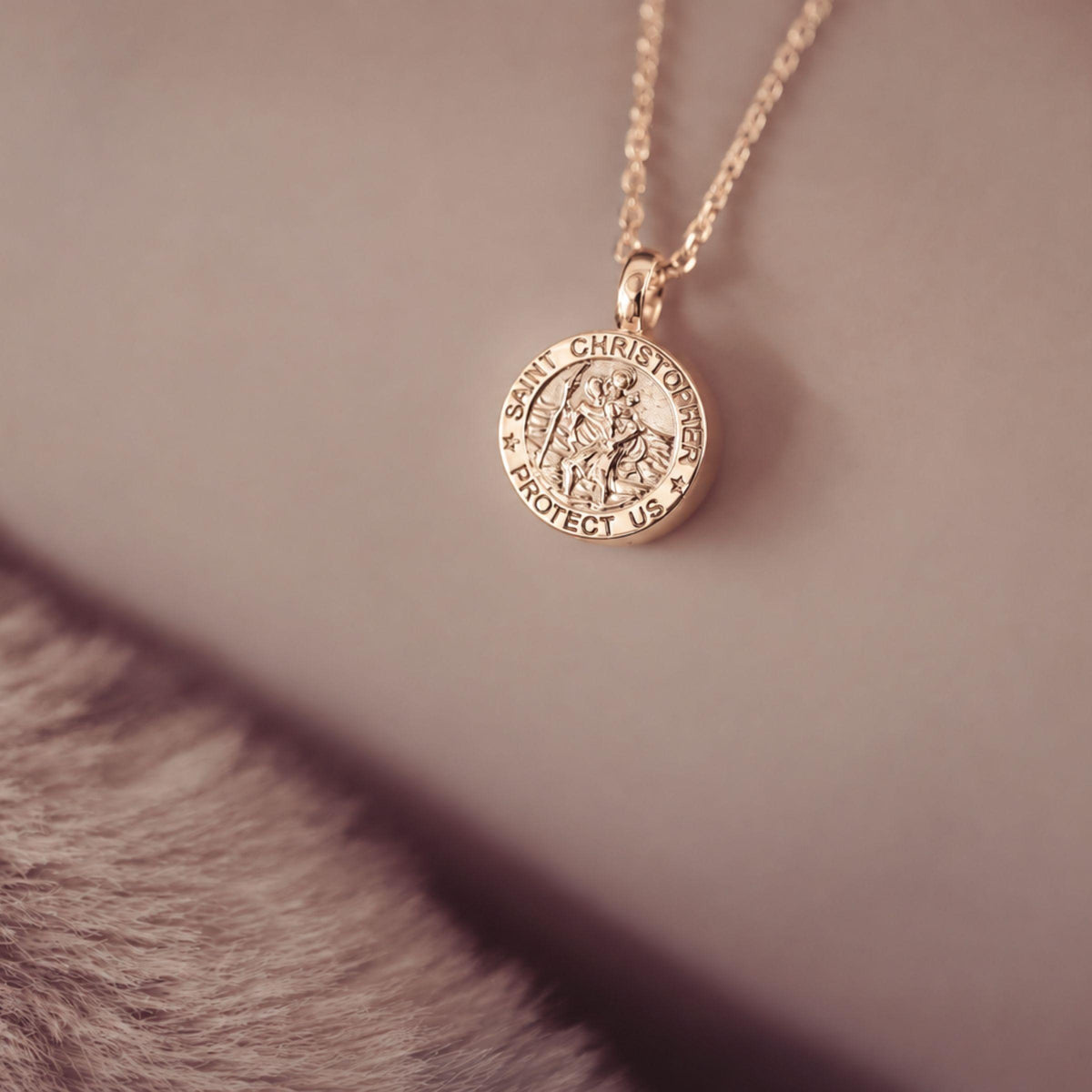 A close-up of a gold Saint Christopher pendant necklace resting on a neutral surface with a soft, out-of-focus textured prop in the foreground.