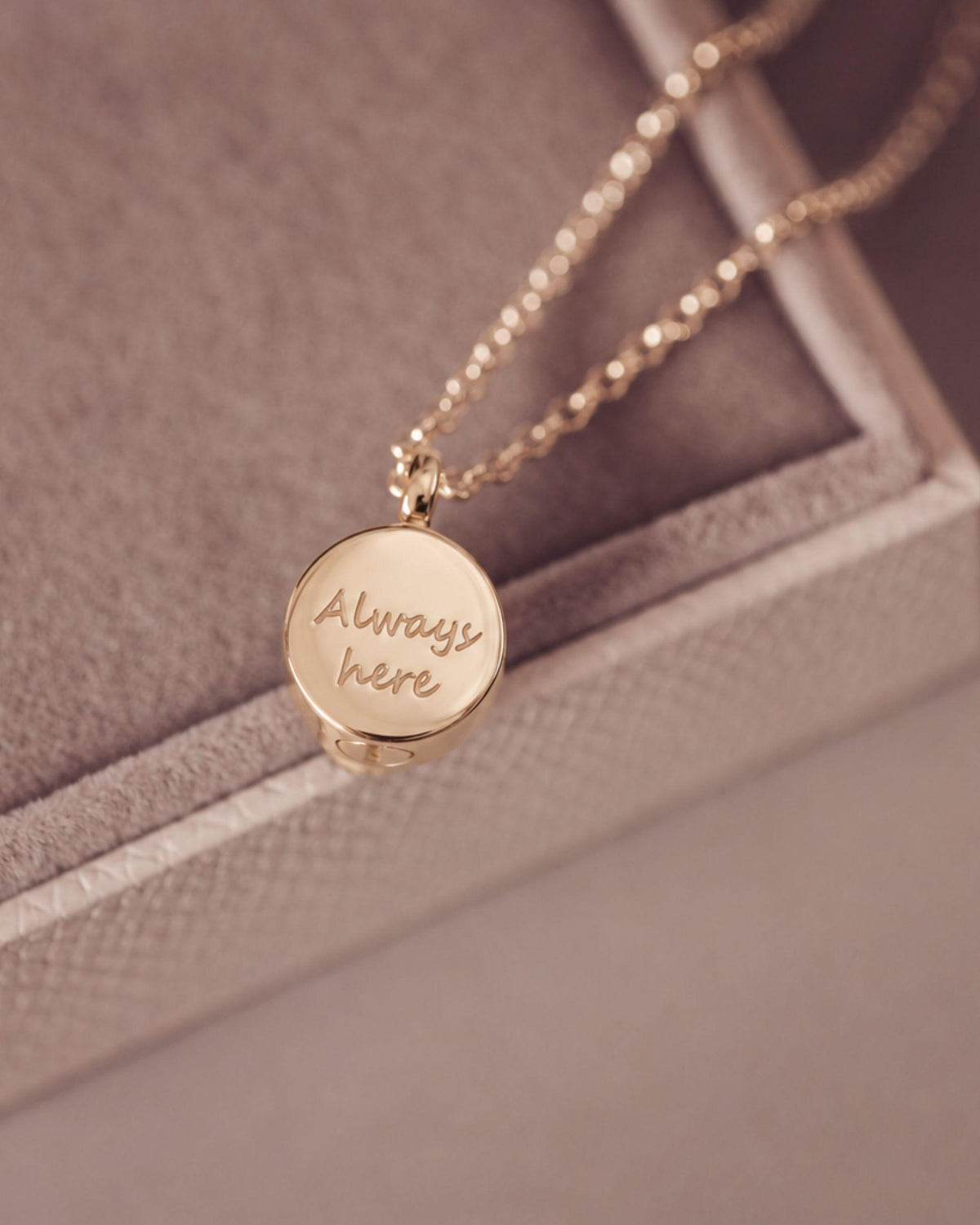 A gold round pendant necklace engraved with the words 'Always here' in script, resting on a textured beige jewelry box.