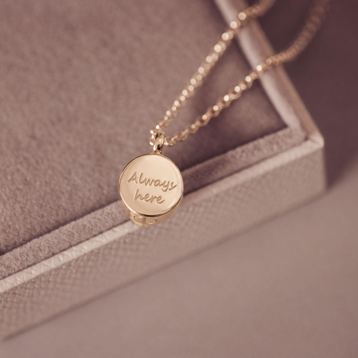 A gold round pendant necklace engraved with the words 'Always here' in script, resting on a textured beige jewelry box.