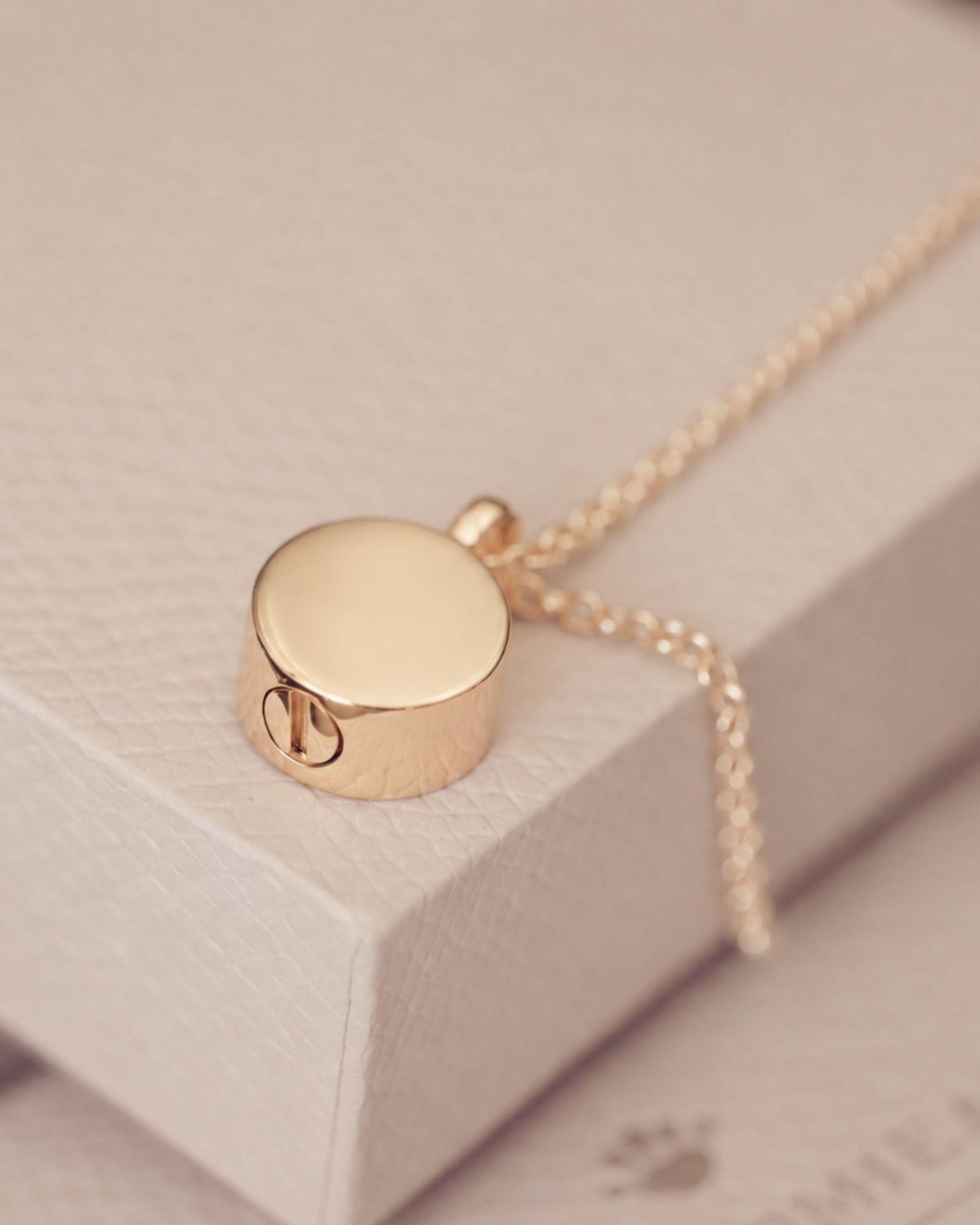 A gold-toned cremation round urn pendant necklace resting on a textured cream-colored jewelry box.
