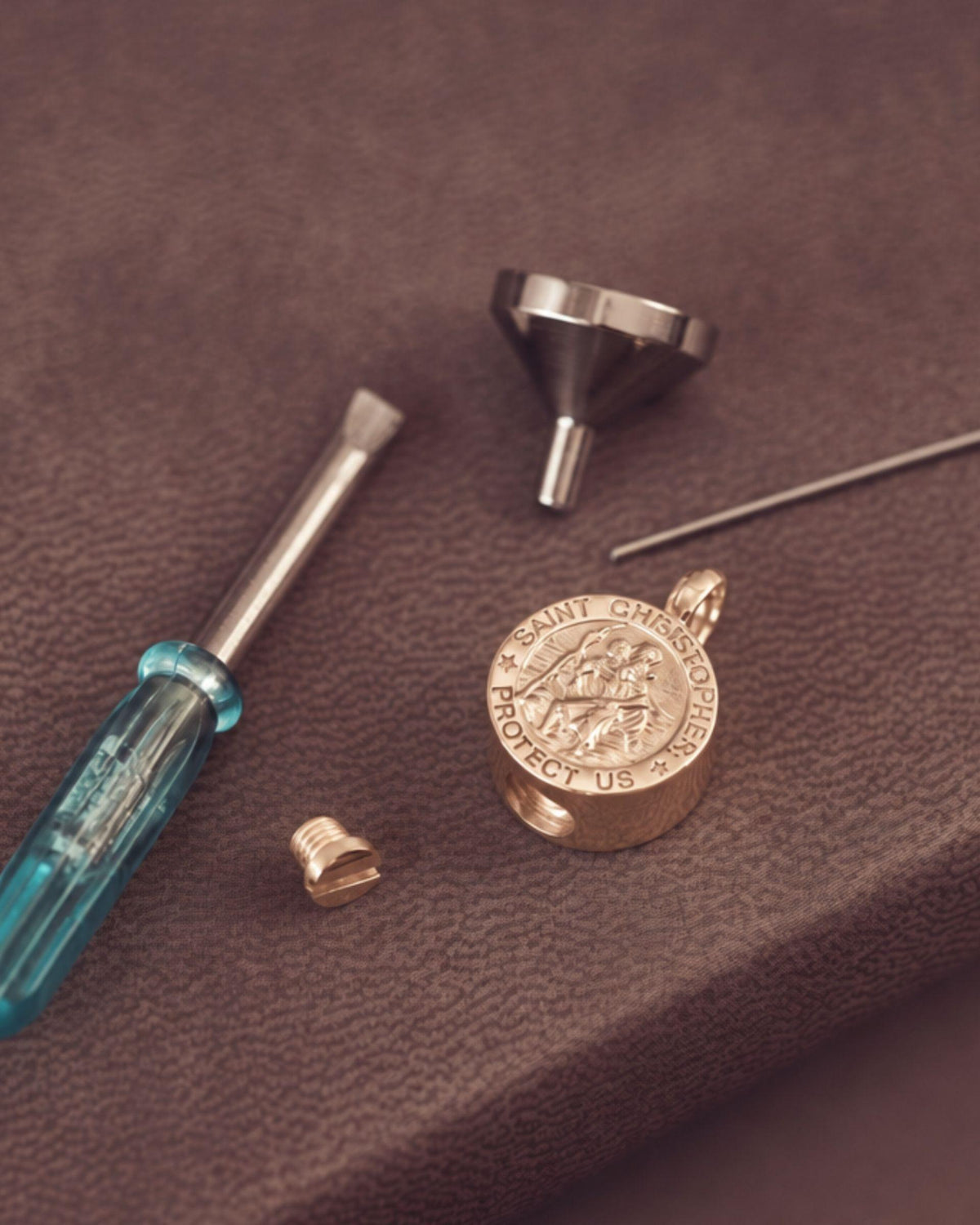 A small set of jewellery tools with a blue-handled screwdriver, metal funnel, small screw piece, and thin pin, arranged on a brown textured surface.