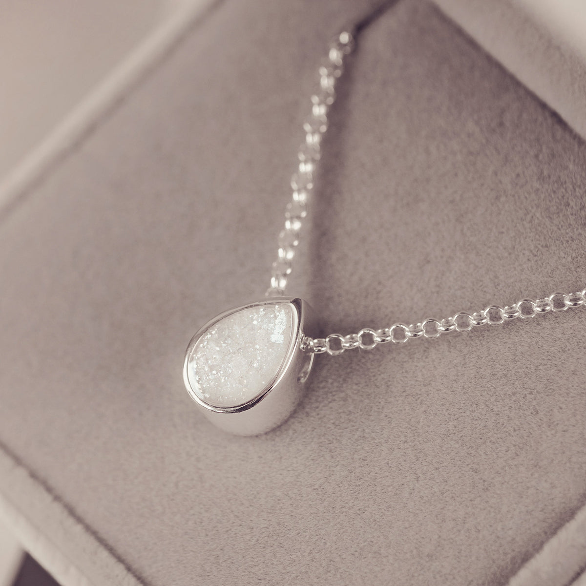 A silver teardrop pendant necklace with a textured, glittery white stone, displayed inside a gray velvet jewelry box.
