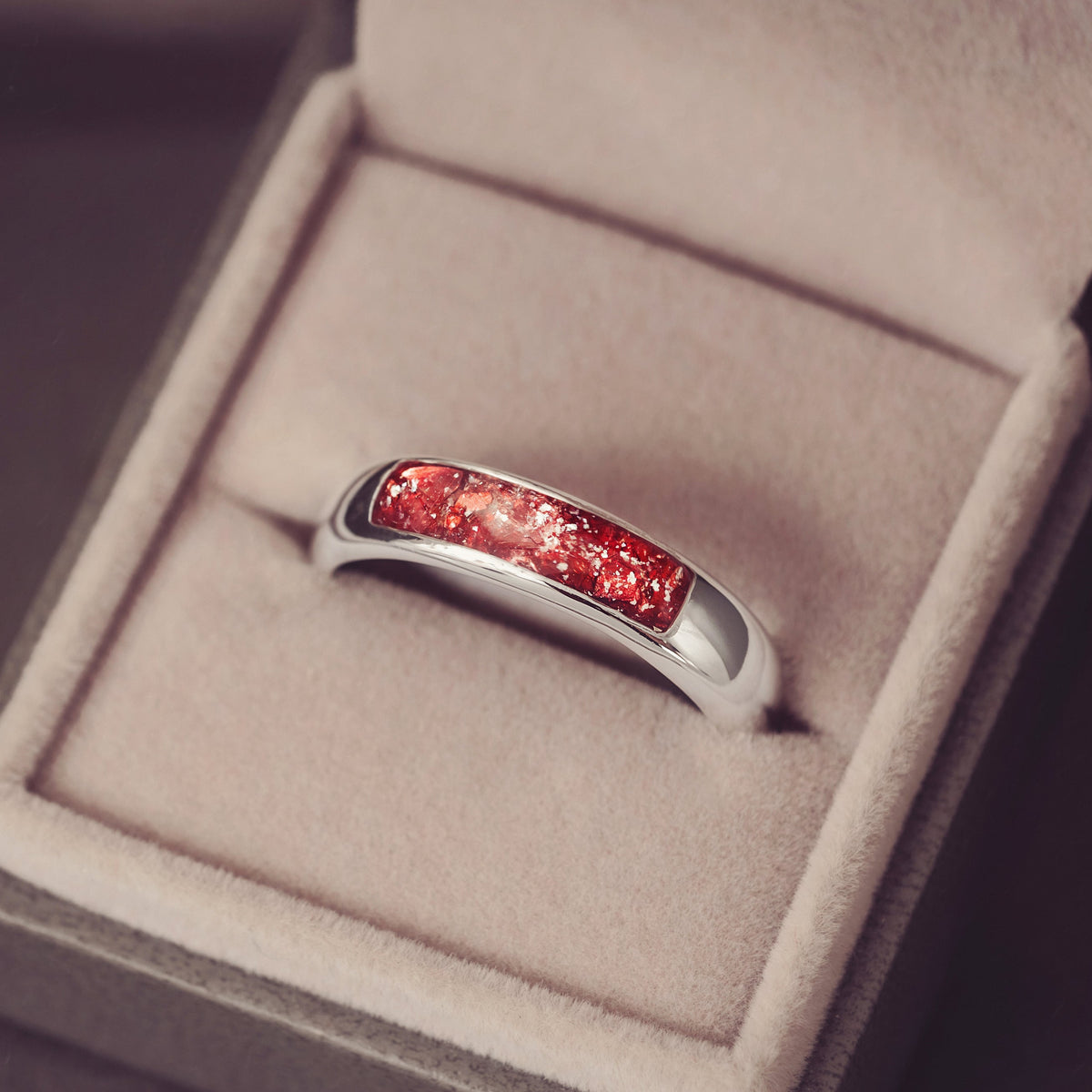 A silver band ring with a curved red inlay, displayed inside an open beige jewellery box on a dark surface.