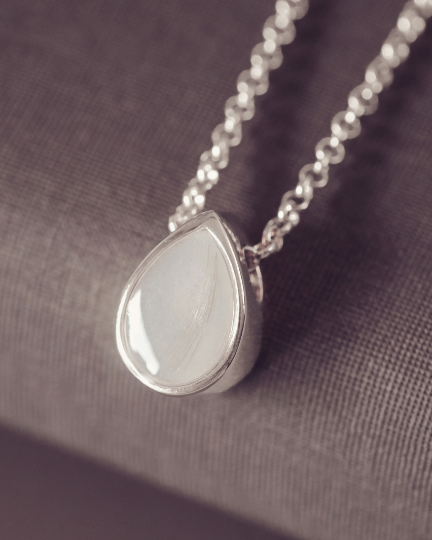 A silver teardrop pendant necklace with a smooth, polished white stone, resting on a textured gray fabric surface.
