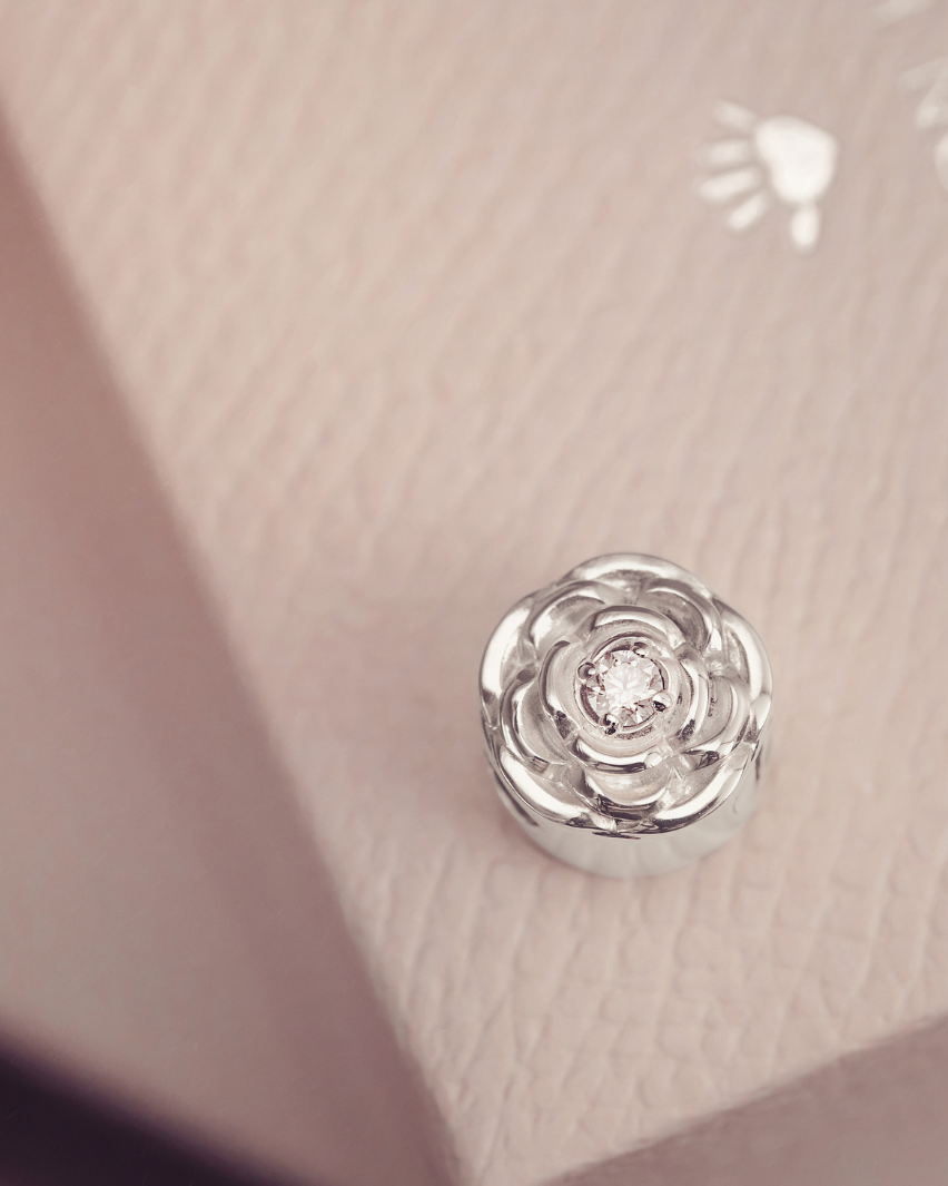 A silver-toned rose-shaped charm with a clear central gemstone, resting on a textured light pink surface.