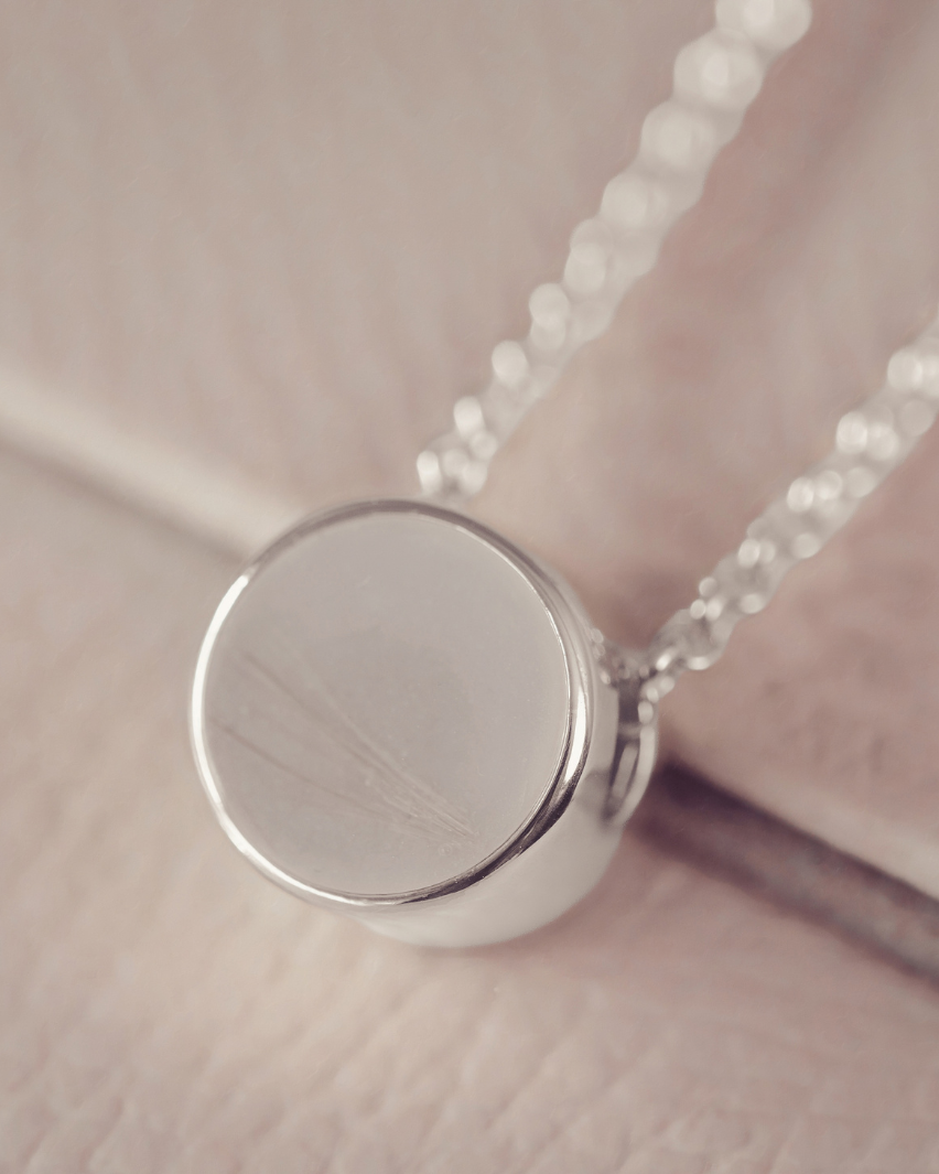 A close-up of a silver pendant necklace with a round, flat disc pendant on a delicate chain, resting on a soft, light pink textured surface. the pendant holds blonde/grey hair