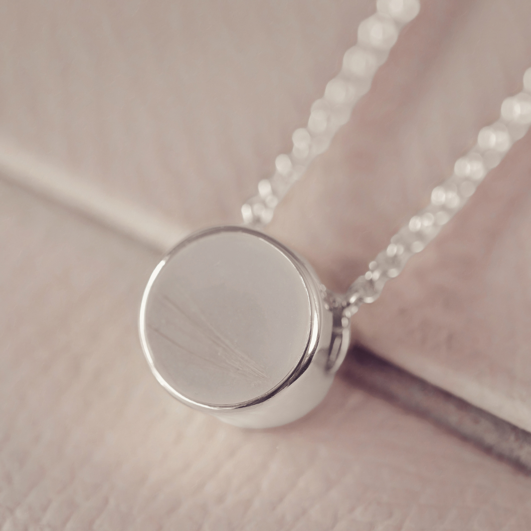 A close-up of a silver pendant necklace with a round, flat disc pendant on a delicate chain, resting on a soft, light pink textured surface. the pendant holds blonde/grey hair