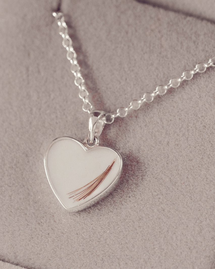 A silver heart-shaped pendant necklace containing a single strand of brown hair, displayed on a soft beige jewelry box.