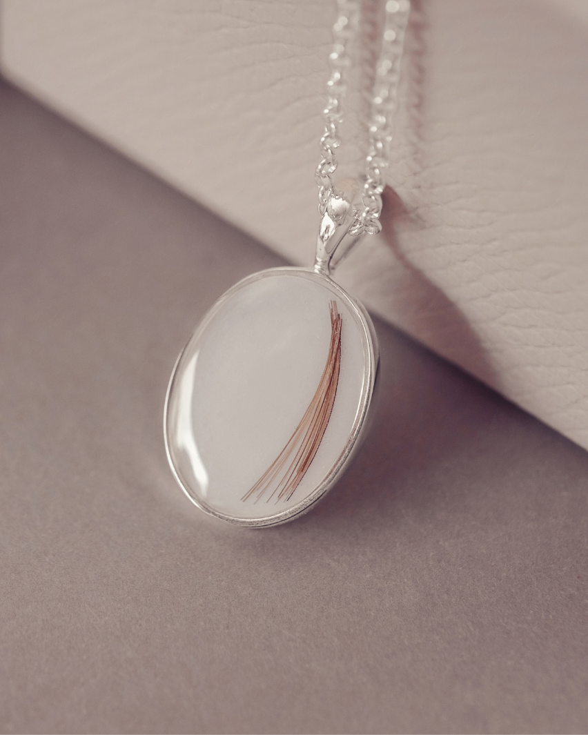 A silver oval pendant necklace containing a single brown hair strand, resting on a neutral gray surface next to a textured cream-colored box.