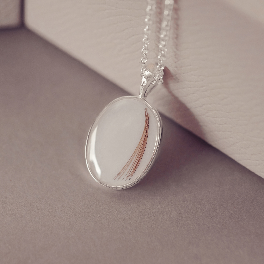 A silver oval pendant necklace containing a single brown hair strand, resting on a neutral gray surface next to a textured cream-colored box.