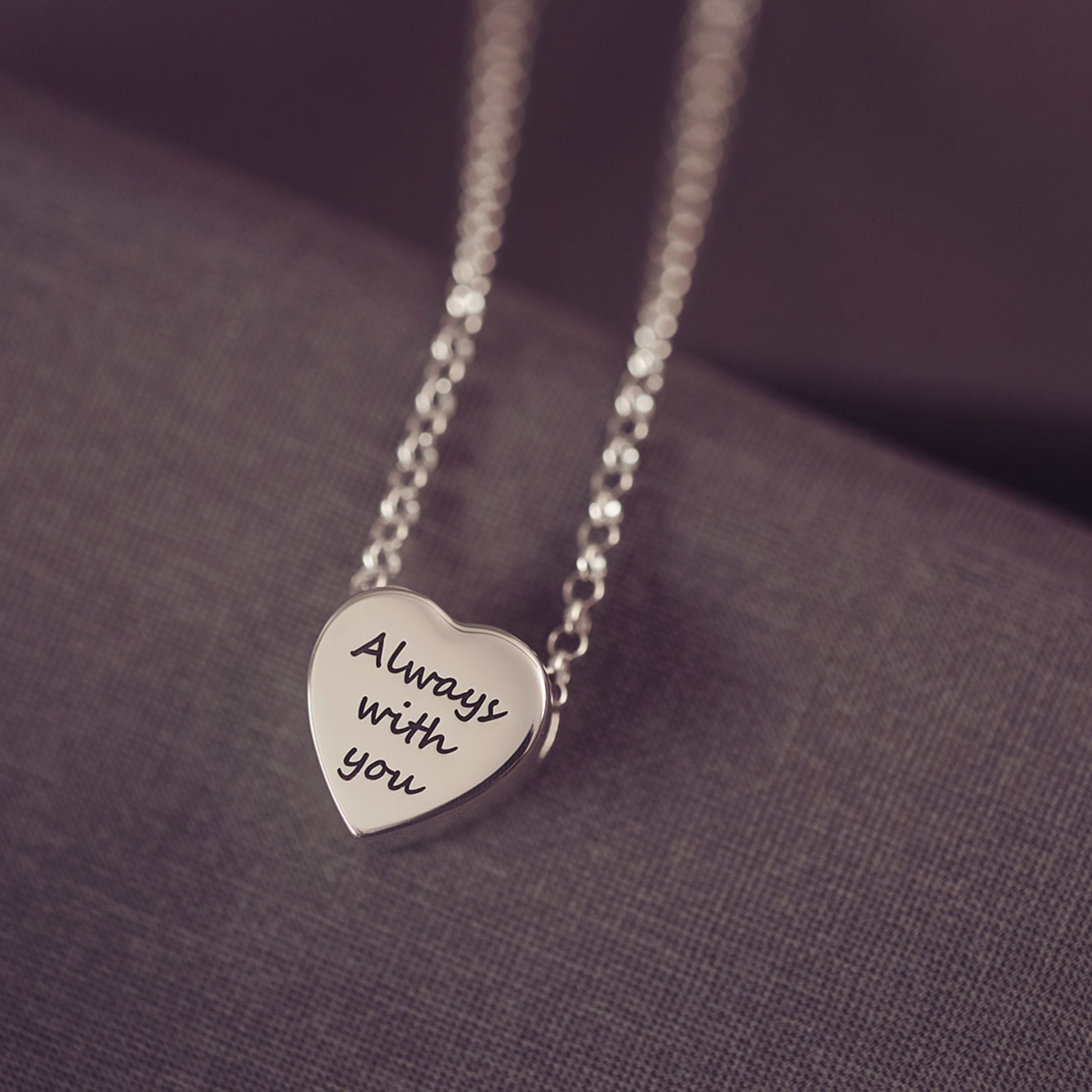 Silver heart-shaped pendant necklace with 'Always with you' engraving on a purple fabric background