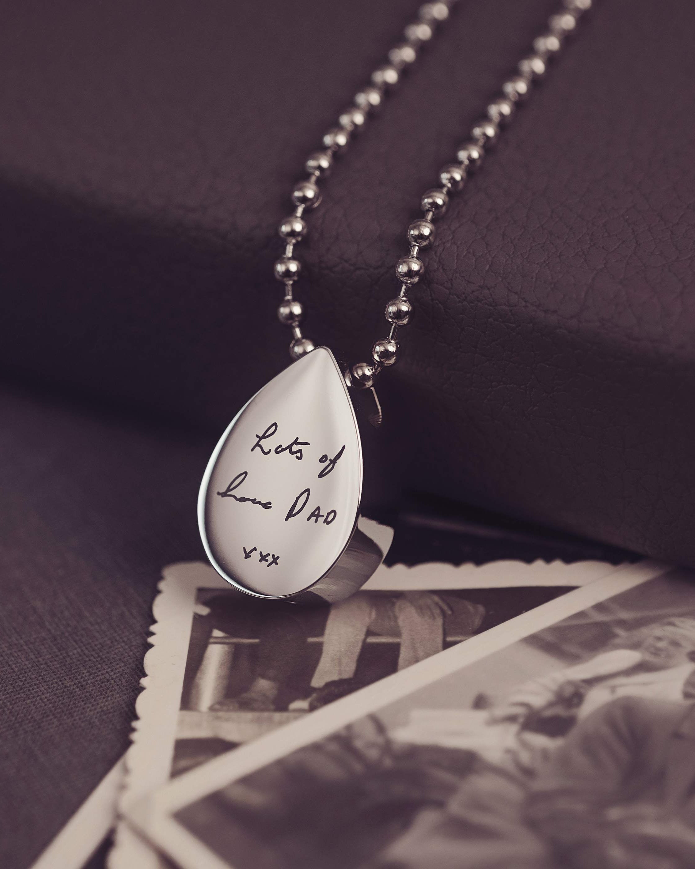 Teardrop-shaped silver pendant with engraved text on a dark background with vintage photos.