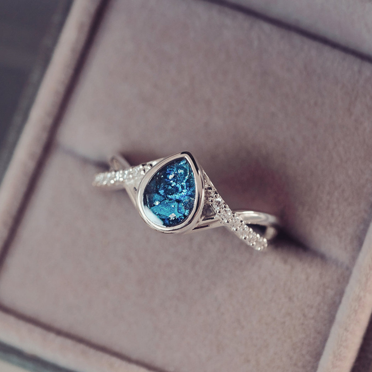 A silver ring with a teardrop-shaped blue gemstone and small clear stones on the band, displayed inside an open graey jewellery box