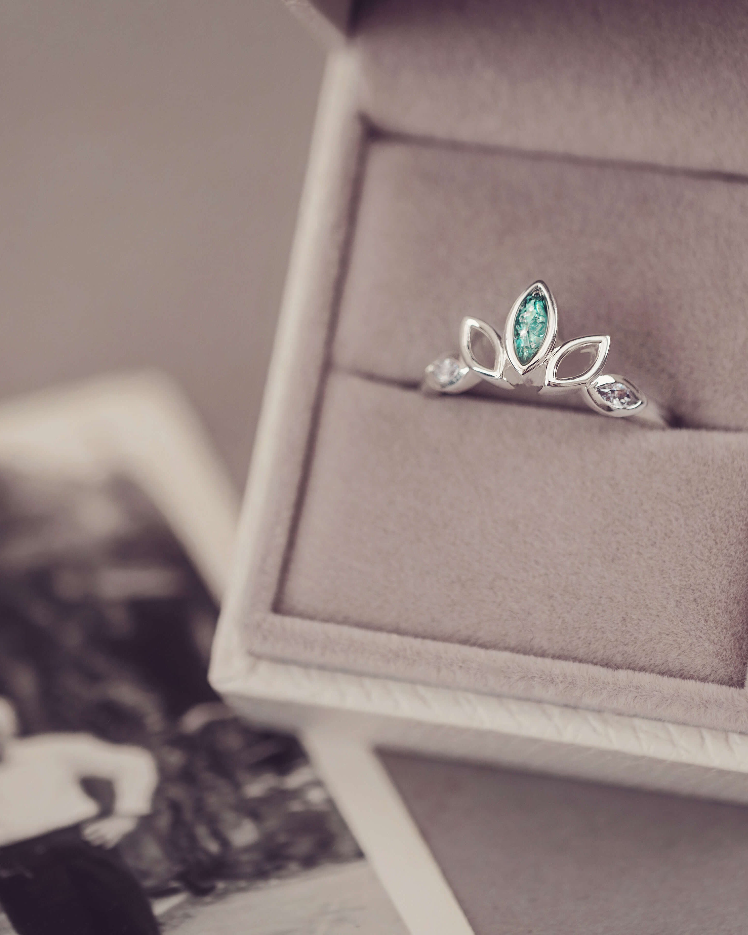 Sterling Silver Ring, created with your loved ones ashes, with Cubic Zircona gemstones. Green floating crystals with in piece in a grey jewellery box placed on top of a blurred photo