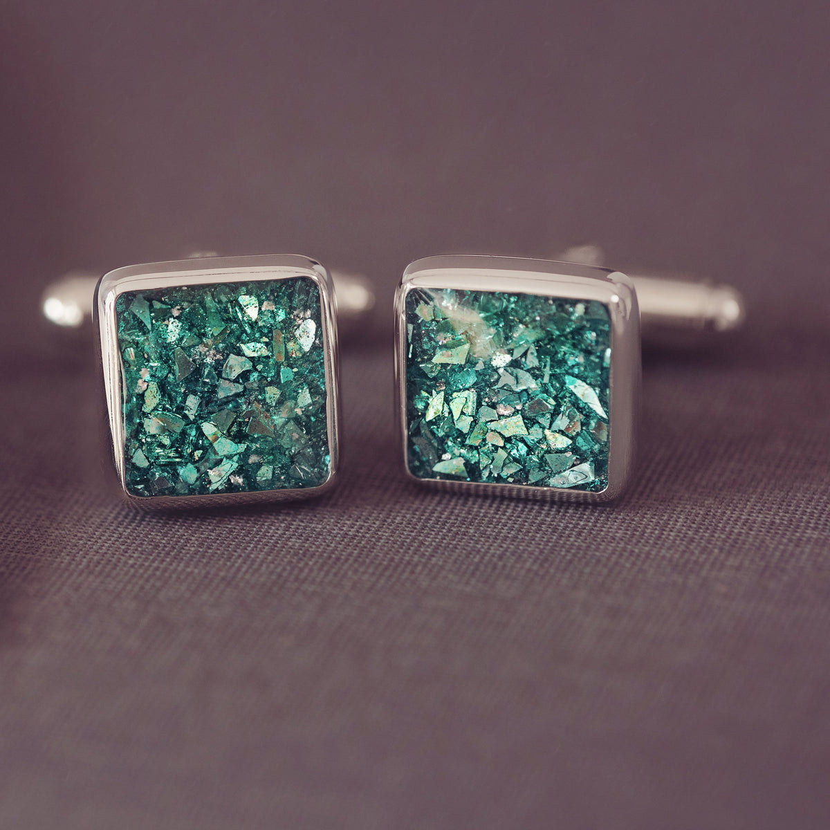 A pair of square silver cufflinks with glittery green and blue mosaic inlays, resting on a textured dark grey surface