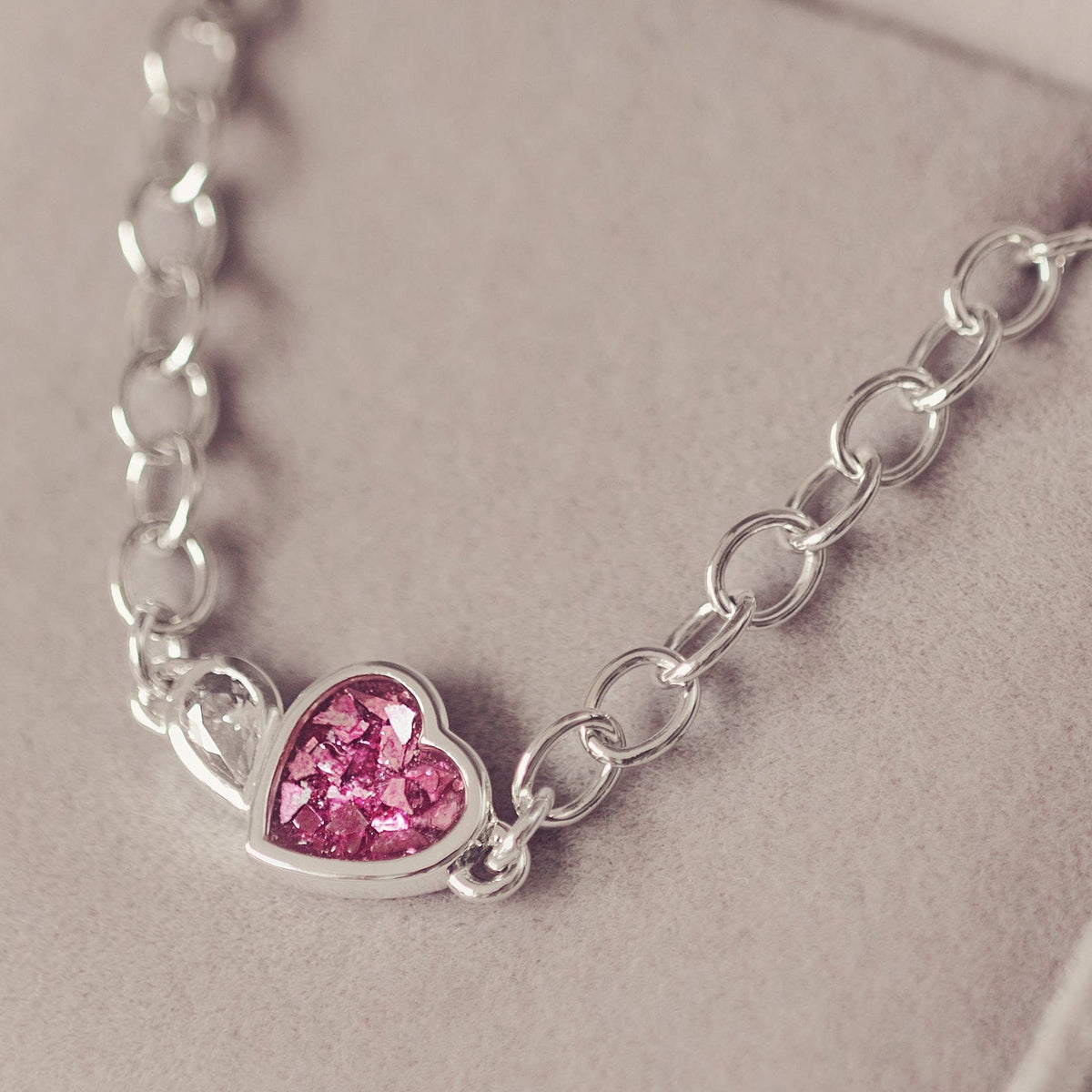 A silver chain necklace with a small heart-shaped pink gemstone pendant, displayed inside an open light grey jewellery box.