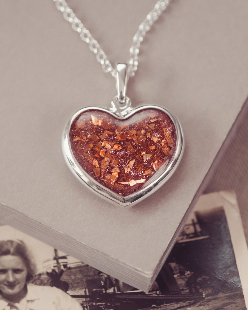 A heart-shaped silver pendant necklace containing shimmering orange-brown glitter, resting on a beige book with an old black-and-white photograph underneath.