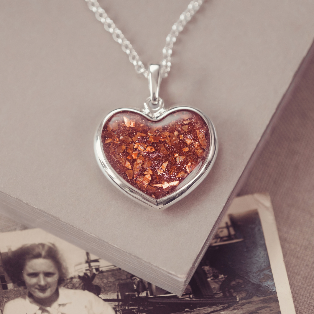 A heart-shaped silver pendant necklace containing shimmering orange-brown glitter, resting on a beige book with an old black-and-white photograph underneath.