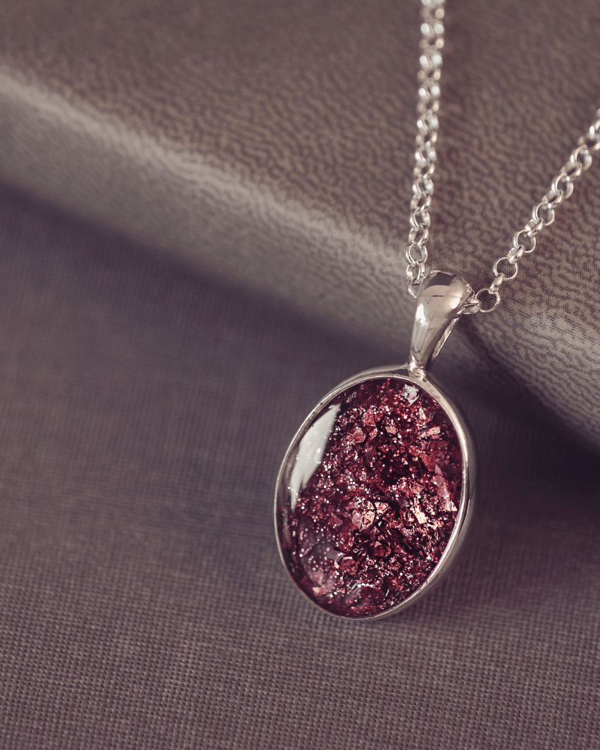 Sterling silver oval necklace with a central section filled with memorial ashes and brown crushed crystals, displayed on a grey jewellery box