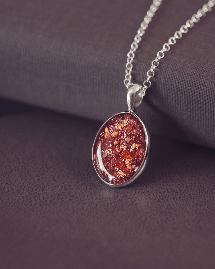 Sterling silver oval necklace with a central section filled with memorial ashes and orange crushed crystals, displayed on a dark background