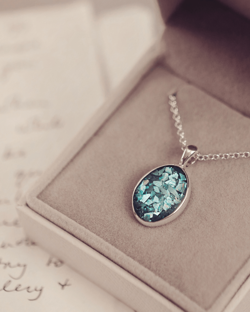 Sterling silver oval necklace with a central section filled with memorial ashes and turquoise crushed crystals, displayed on a grey fabric background