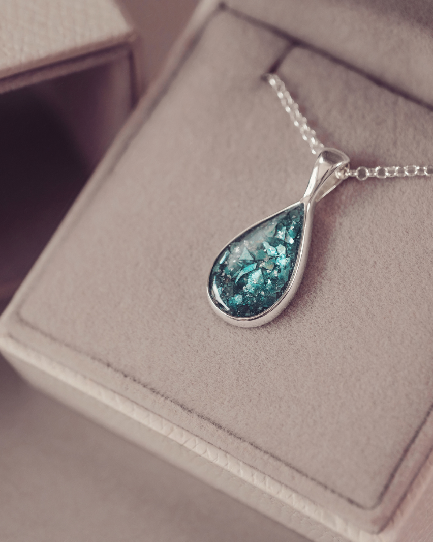 A silver teardrop pendant necklace with a textured, glittery teal-blue center, resting on a beige jewelry box.