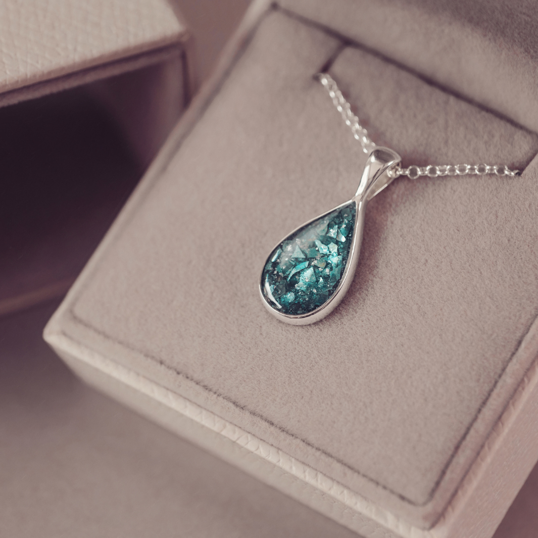 A silver teardrop pendant necklace with a textured, glittery teal-blue center, resting on a beige jewelry box.