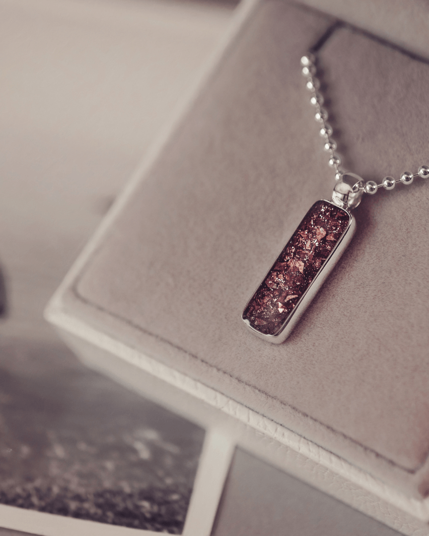 A sophisticated Mens silver necklace featuring a rectangular pendant filled with shimmering brown crystal and resin, delicately layered with cremation ashes, showcased against a dark background on a silver ball chain