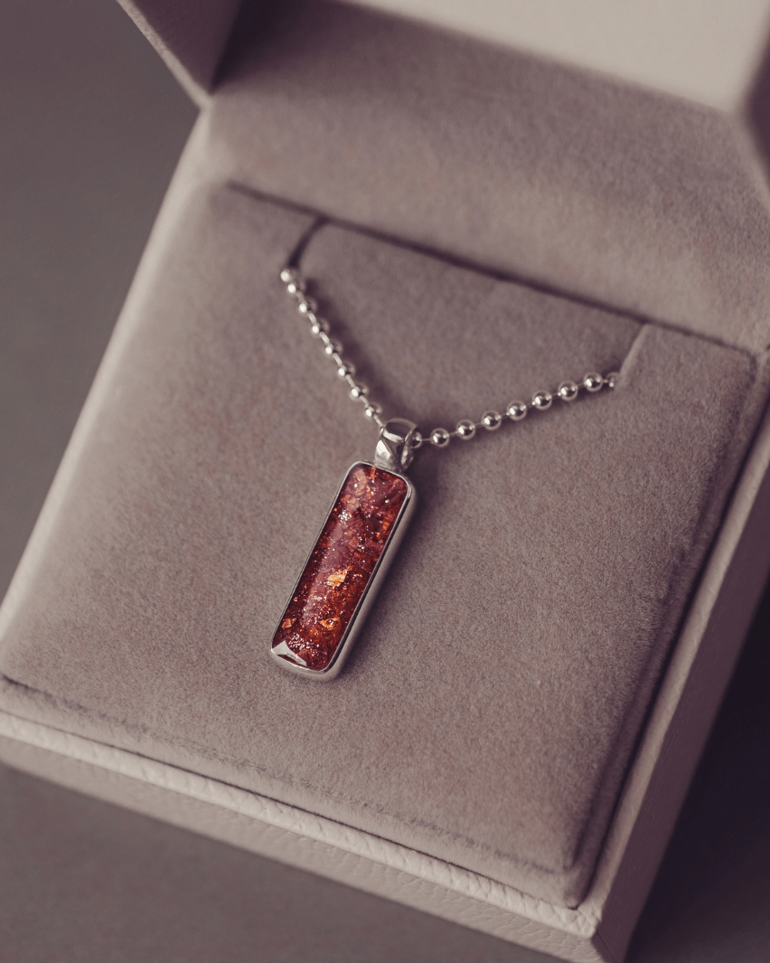A sophisticated Mens silver necklace featuring a rectangular pendant filled with shimmering orange crystal and resin, delicately layered with cremation ashes, showcased against a dark background on a silver ball chain