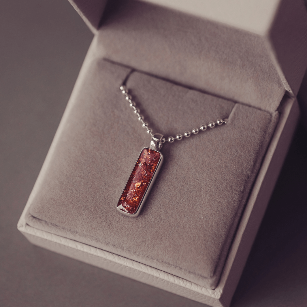 A sophisticated Mens silver necklace featuring a rectangular pendant filled with shimmering orange crystal and resin, delicately layered with cremation ashes, showcased against a dark background on a silver ball chain
