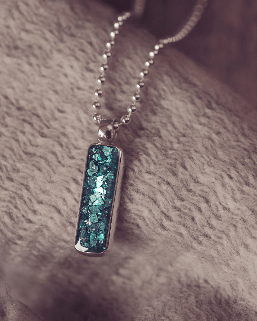 A sophisticated Mens silver necklace featuring a rectangular pendant filled with shimmering turquoise crystal and resin, delicately layered with cremation ashes, showcased against a textured background on a silver ball chain