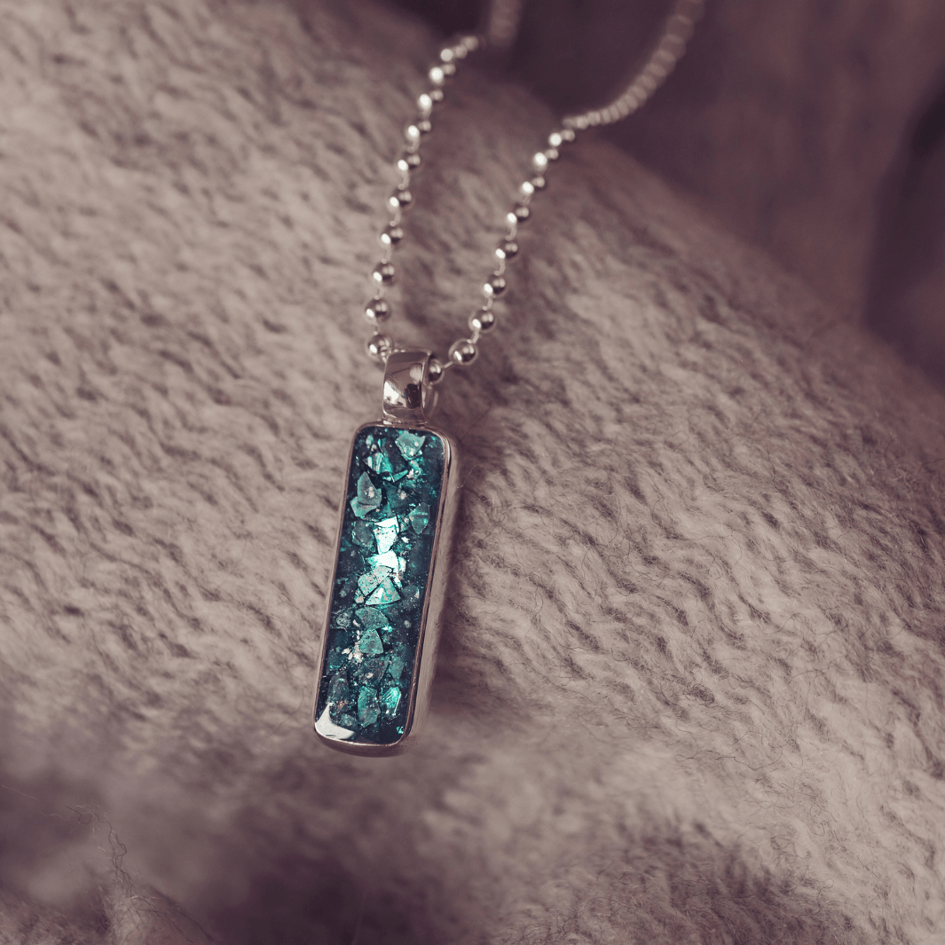 A sophisticated Mens silver necklace featuring a rectangular pendant filled with shimmering turquoise crystal and resin, delicately layered with cremation ashes, showcased against a textured background on a silver ball chain