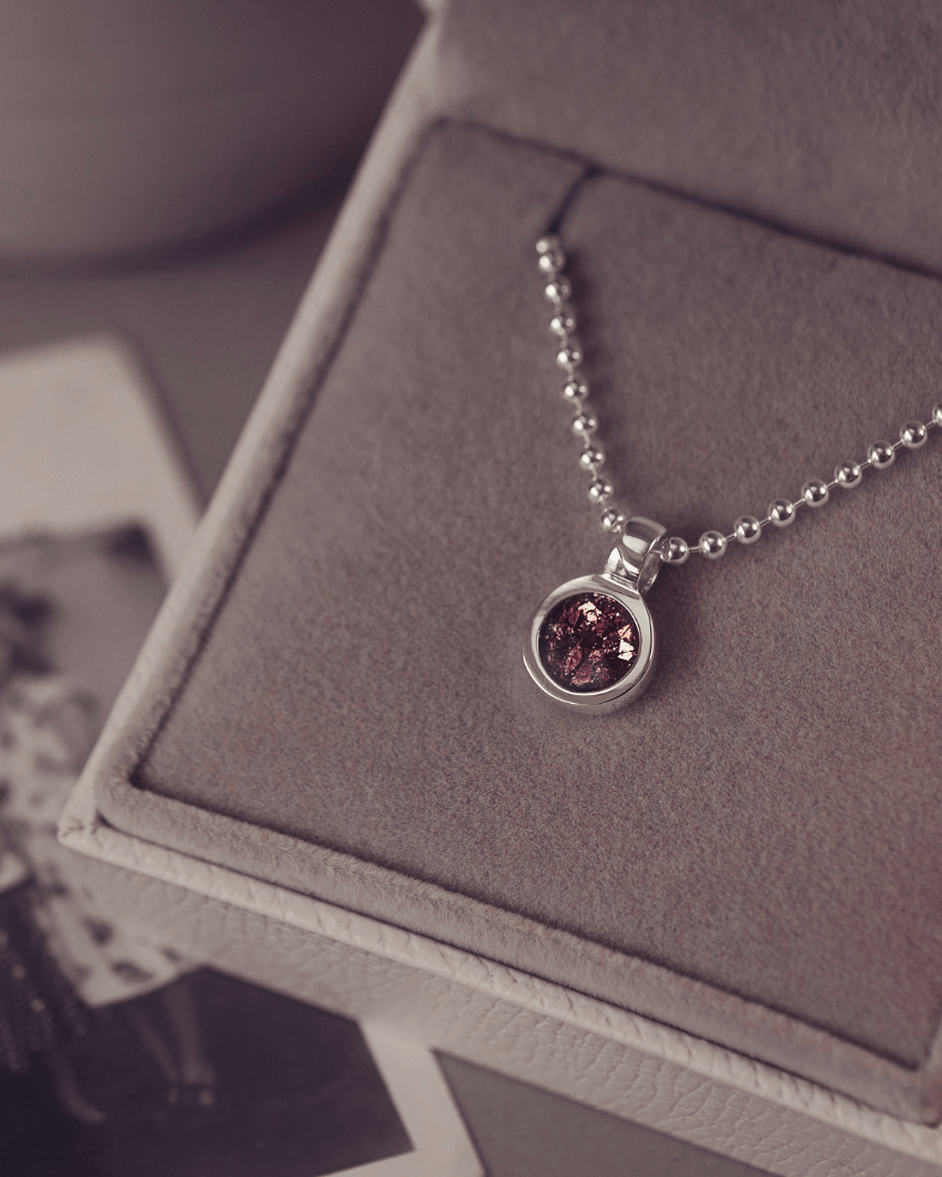 A sophisticated Mens silver necklace featuring a small round pendant filled with shimmering brown crystal and resin, delicately layered with cremation ashes, showcased against a dark background on a silver ball chain
