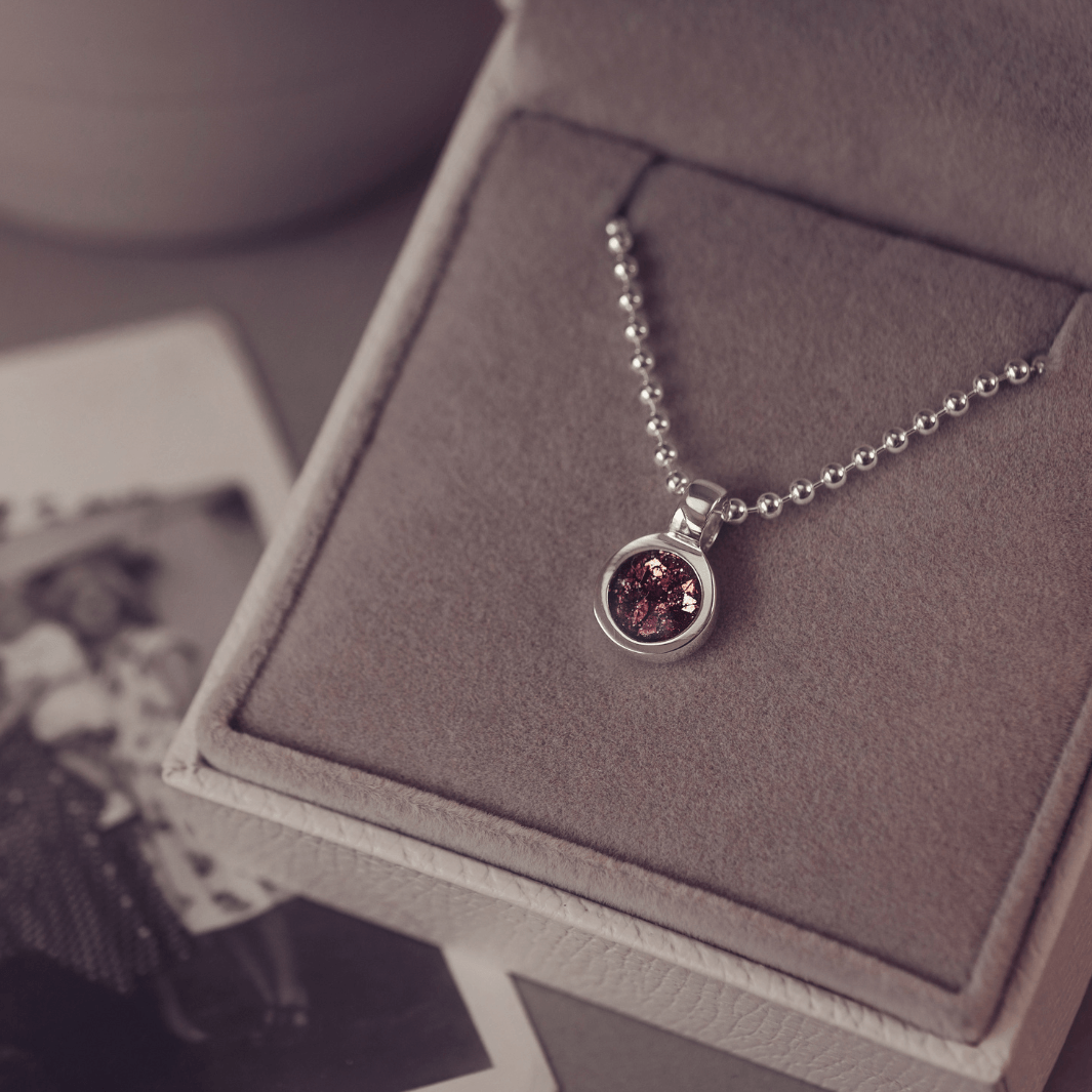 A sophisticated Mens silver necklace featuring a small round pendant filled with shimmering brown crystal and resin, delicately layered with cremation ashes, showcased against a dark background on a silver ball chain