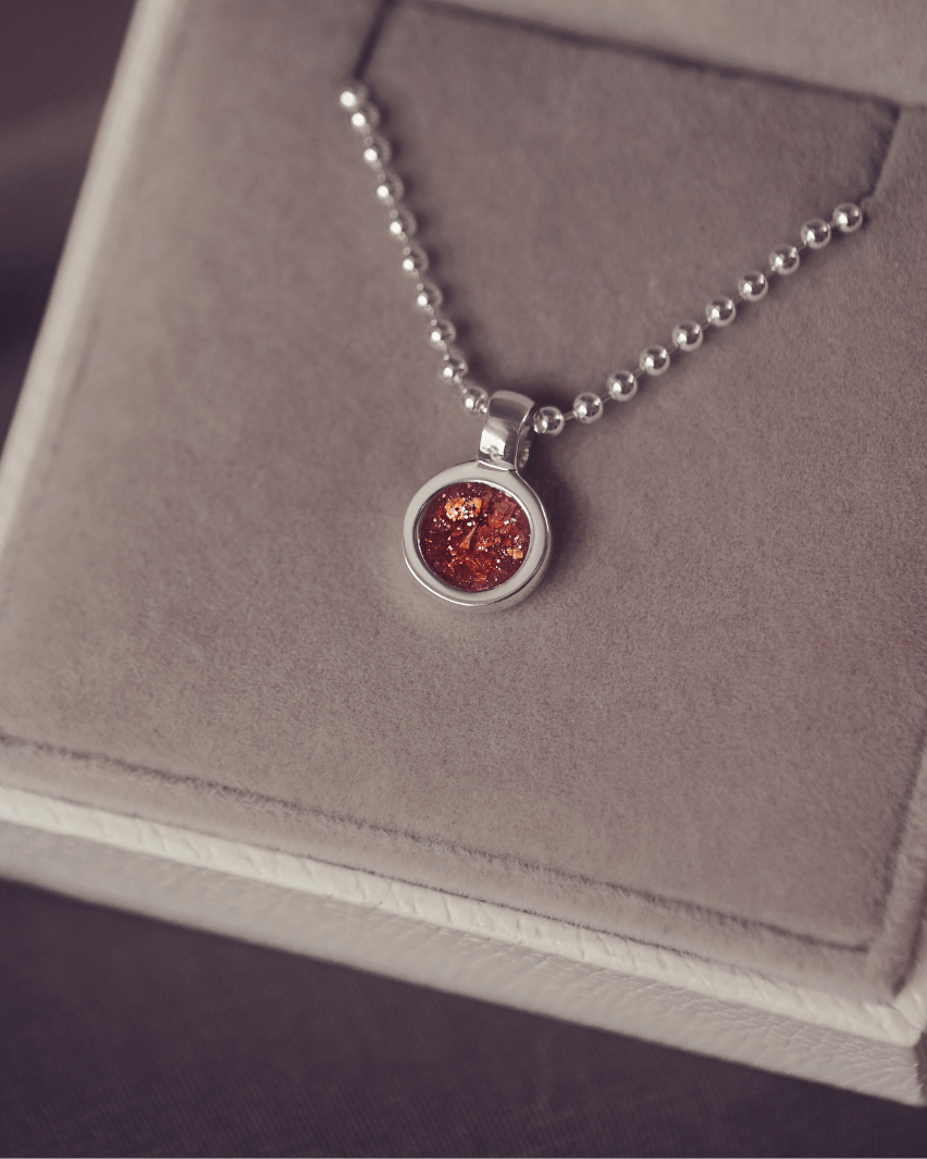 A sophisticated Mens silver necklace featuring a small round pendant filled with shimmering orange crystal and resin, delicately layered with cremation ashes, showcased against a dark background on a silver ball chain