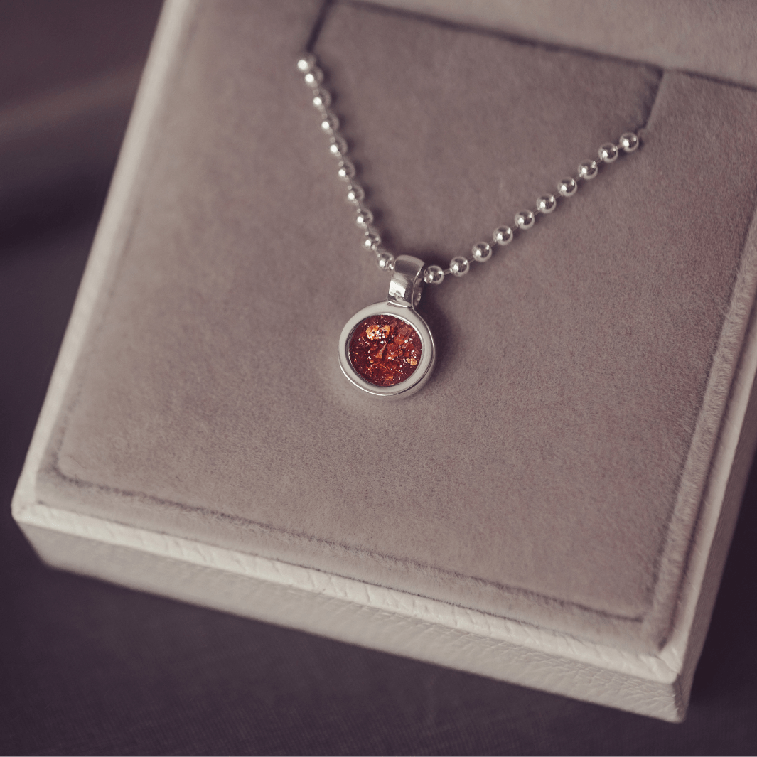 A sophisticated Mens silver necklace featuring a small round pendant filled with shimmering orange crystal and resin, delicately layered with cremation ashes, showcased against a dark background on a silver ball chain