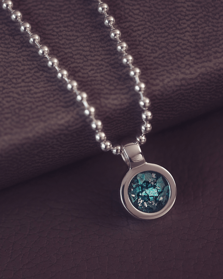 A sophisticated Mens silver necklace featuring a small round pendant filled with shimmering turquoise crystal and resin, delicately layered with cremation ashes, showcased against a dark background on a silver ball chain
