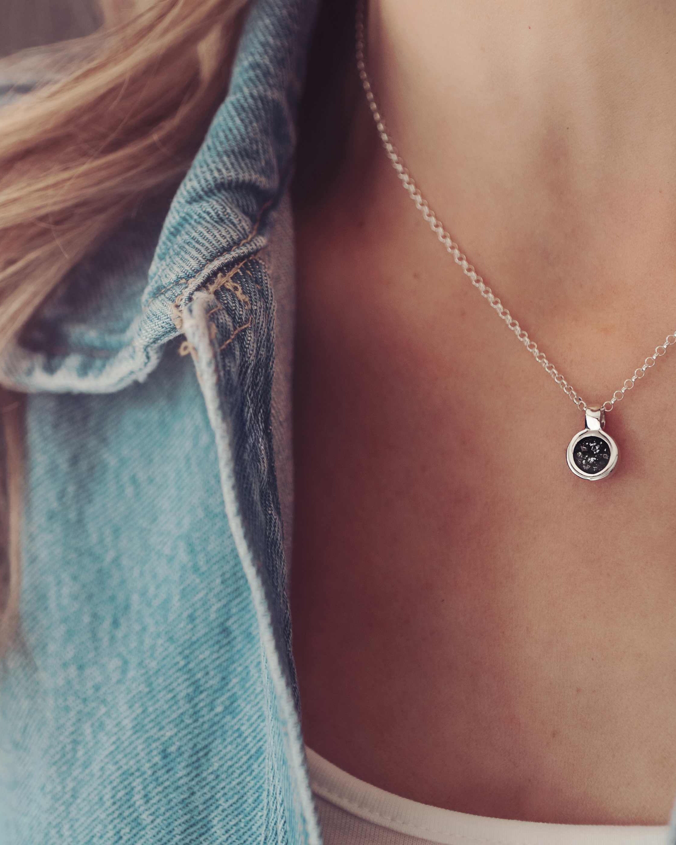 Lady wearing a small round cremation ashes necklace in the colour black. Lady wears a blue denim jacket.