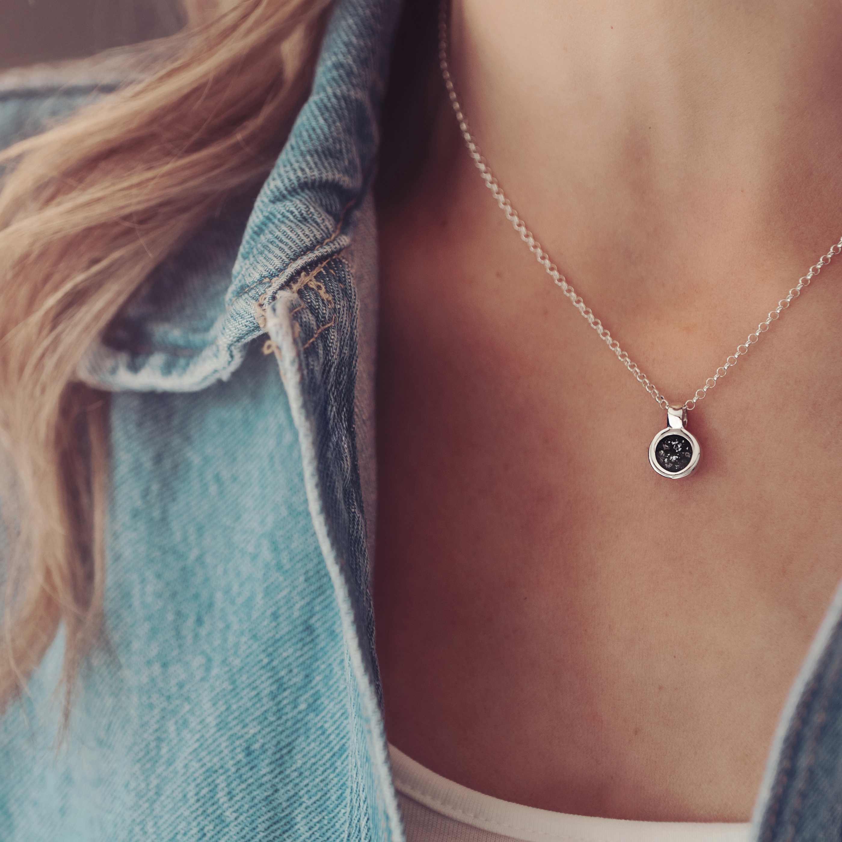 Lady wearing a small round cremation ashes necklace in the colour black. Lady wears a blue denim jacket.