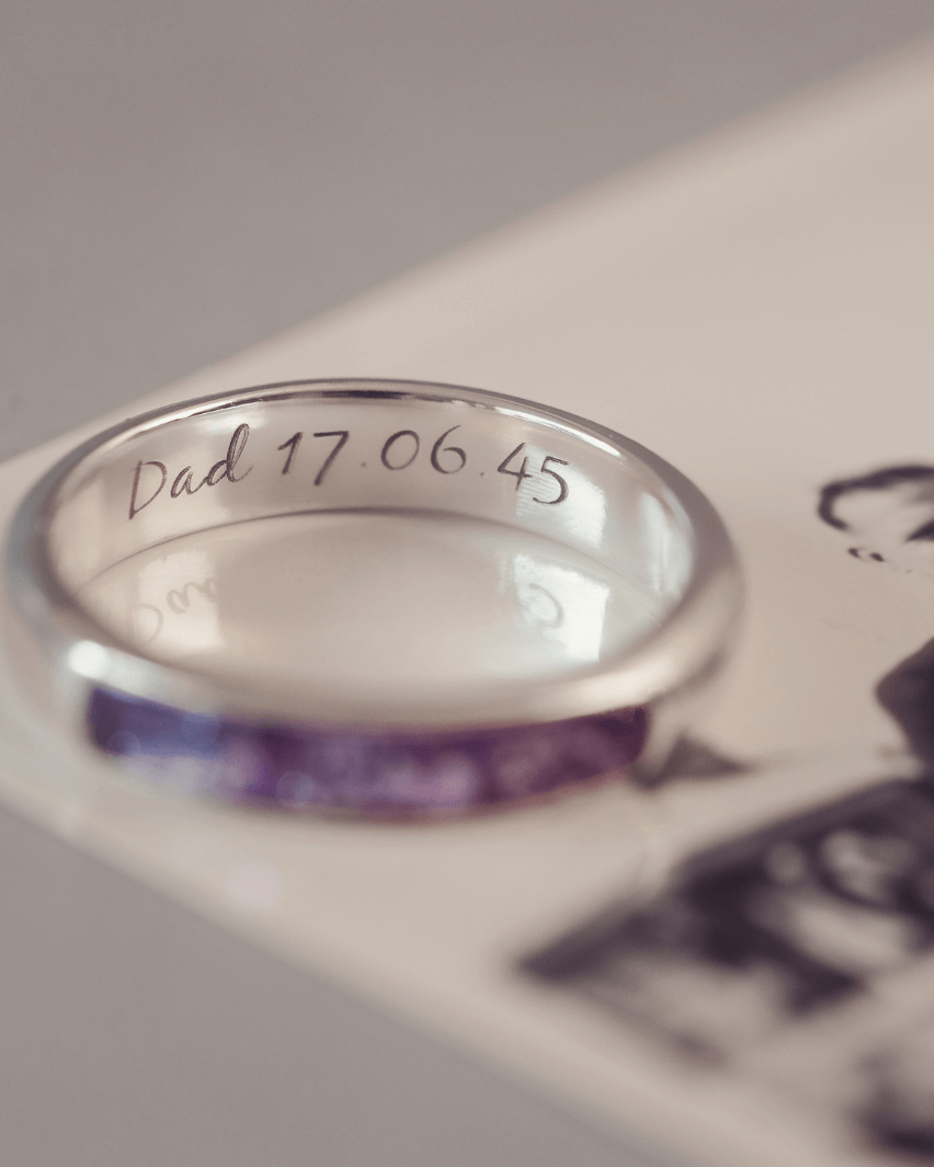 Sterling silver womens ring band with a central section filled with  memorial ashes and purple crushed crystals, on a blurred pale background. The ring has the message 'DAD 17.06.45' engraved into the inside of the ring band in black