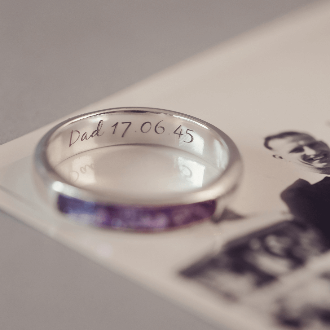 Sterling silver womens ring band with a central section filled with  memorial ashes and purple crushed crystals, on a blurred pale background. The ring has the message 'DAD 17.06.45' engraved into the inside of the ring band in black