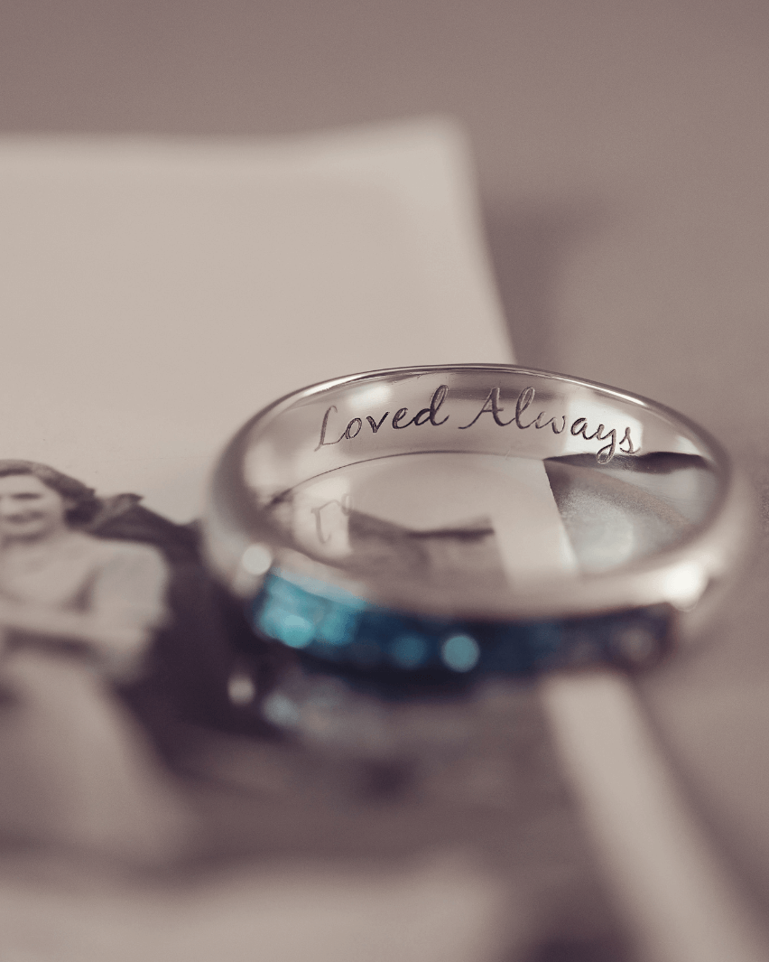 Sterling silver womens ring band with a central section filled with  memorial ashes and blue crushed crystals, on a blurred pale background. The ring has the message 'Loved Always' engraved into the inside of the ring band in black