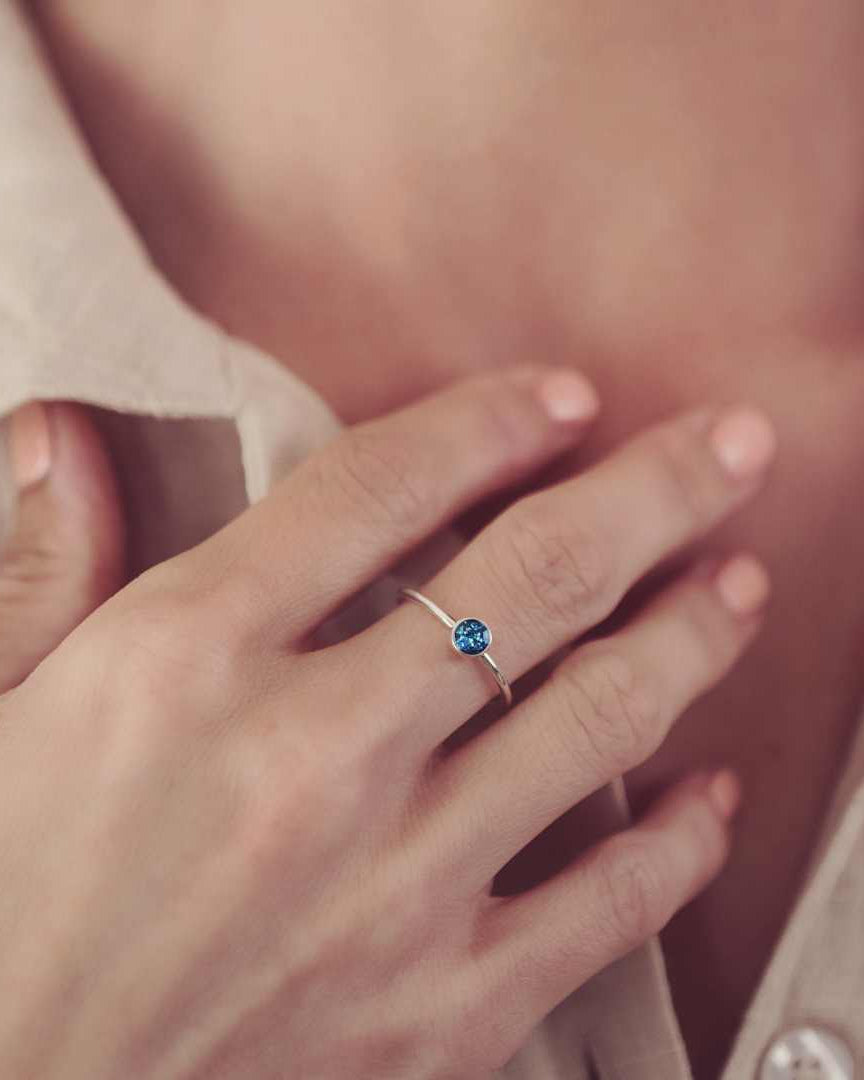 A small round ashes ring in a blue colour. The ring is worn on a woman's finger with her hand to her chest. You can see her white shirt in the background.