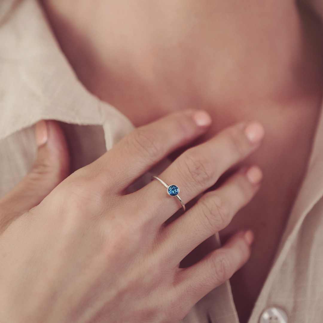 A small round ashes ring in a blue colour. The ring is worn on a woman's finger with her hand to her chest. You can see her white shirt in the background.