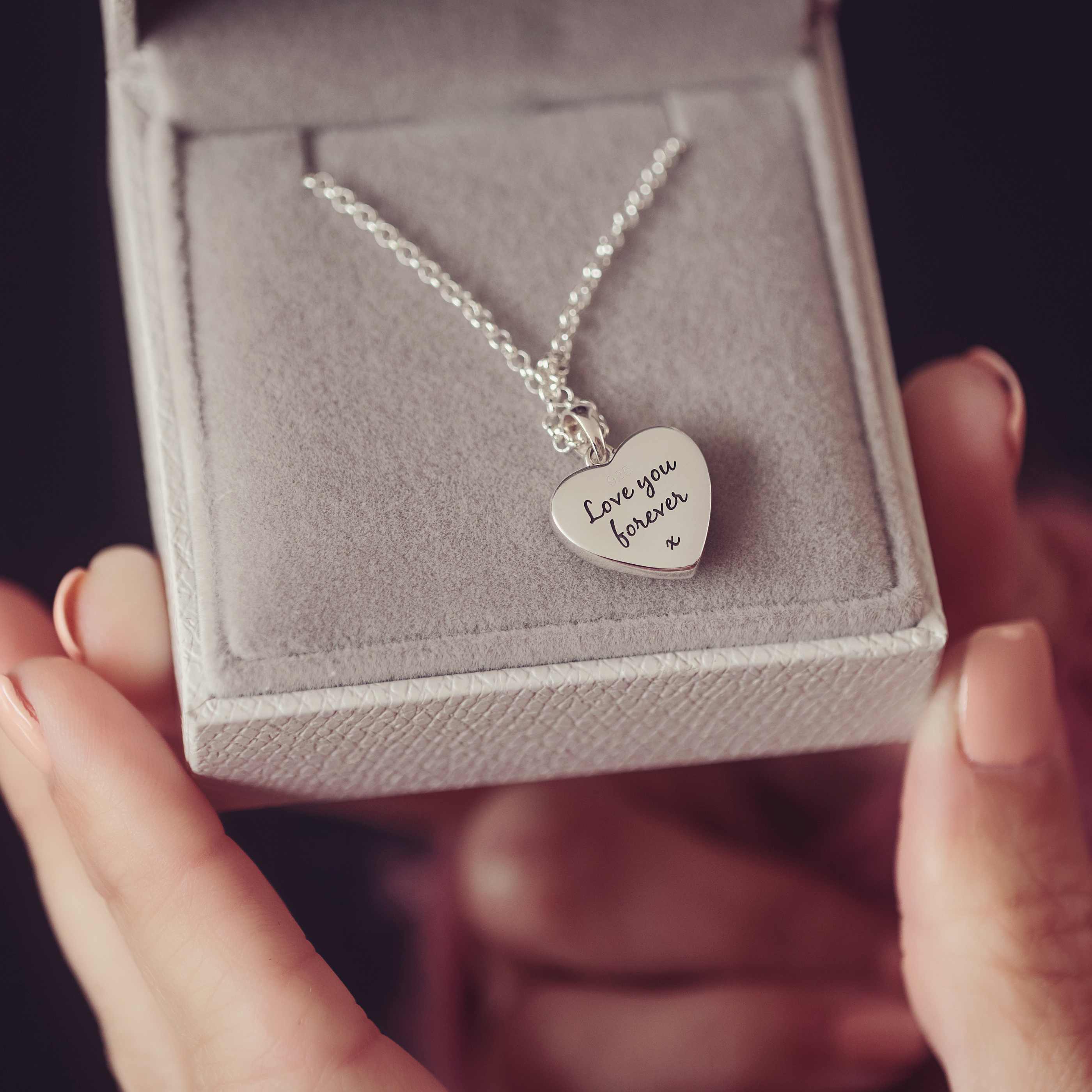 A close up shot of a lady's hands holding a grey jewellery box. Inside the box is a silver heart necklace with engraving.