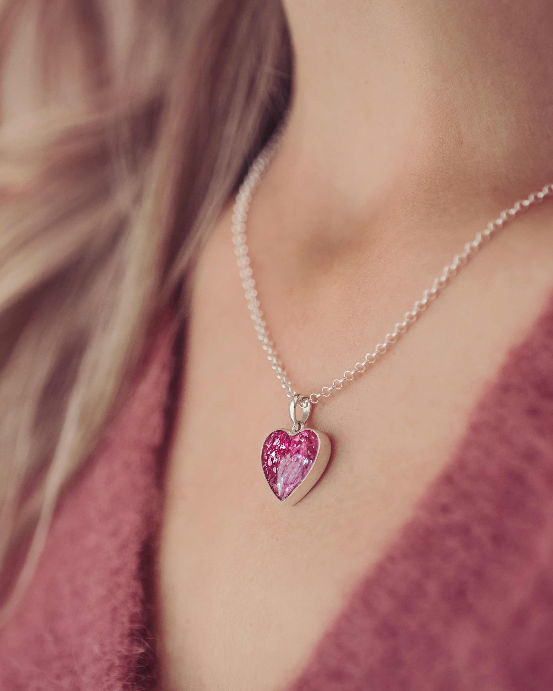 image cropped into a lady's neck showing a small silver and pink heart necklace. The necklace is filled with cremation ashes.